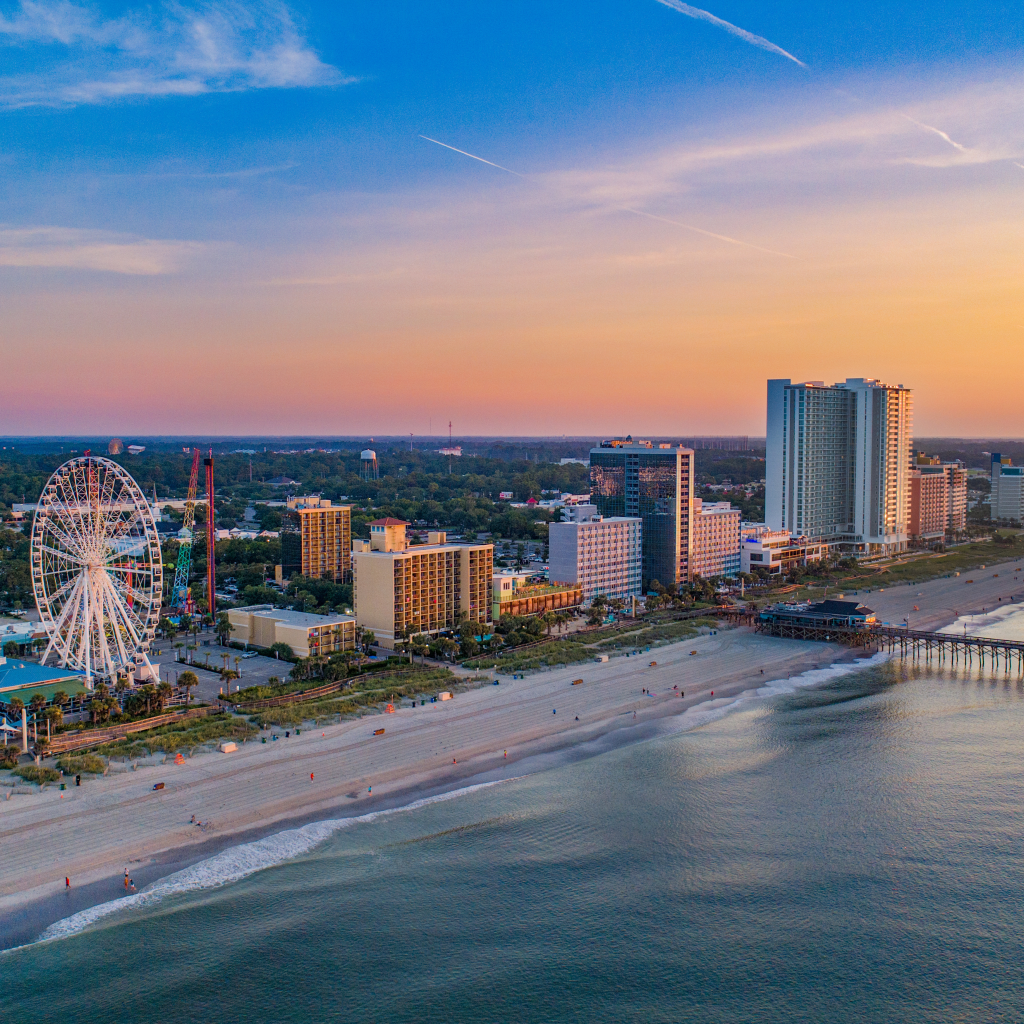 Sunset along the coast showing hotel buildings, a pier, and the Sky Wheel in Myrtle Beach, South Carolina.