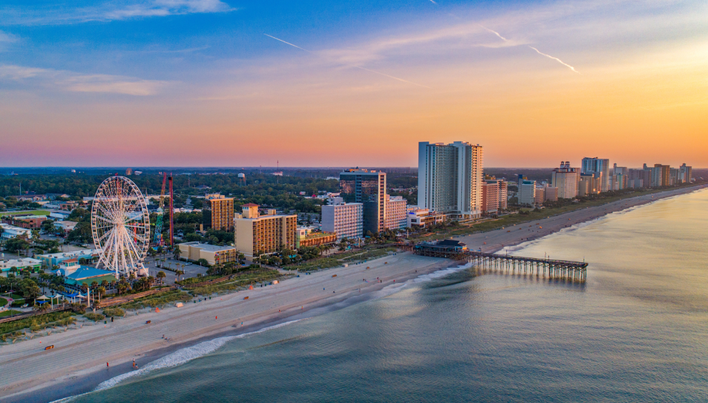 Sunset along the coast showing hotel buildings, a pier, and the Sky Wheel in Myrtle Beach, South Carolina.